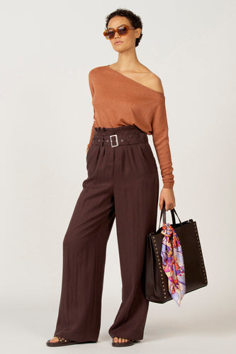 Vrouw in koperkleurige top en bruine pantalon met riem, draagt een zwarte tas met sjaal en sandalen, tegen een witte achtergrond.