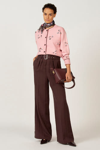 Gilet rose d'Astrid Black Label, avec fermeture à boutons et applications décoratives, associé à un pantalon marron, une ceinture et un sac.
