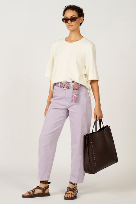 Vrouw in lila broek, wit shirt, bloemenriem en donkerbruine tas.