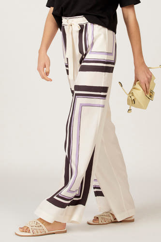 Multikleurige broek van Astrid Black Label, met geometrische print in zwart en lila, gecombineerd met gevlochten sandalen en een gele tas.
