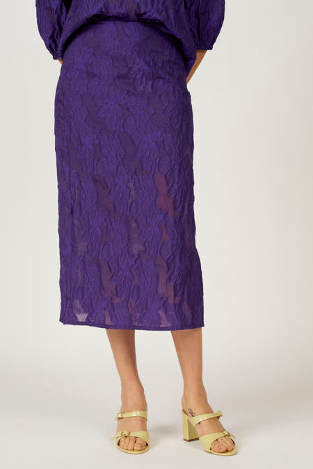 Paarse lange rok van Astrid Black Label, met een bloemmotief en textuur, gecombineerd met gele sandalen met hak.
