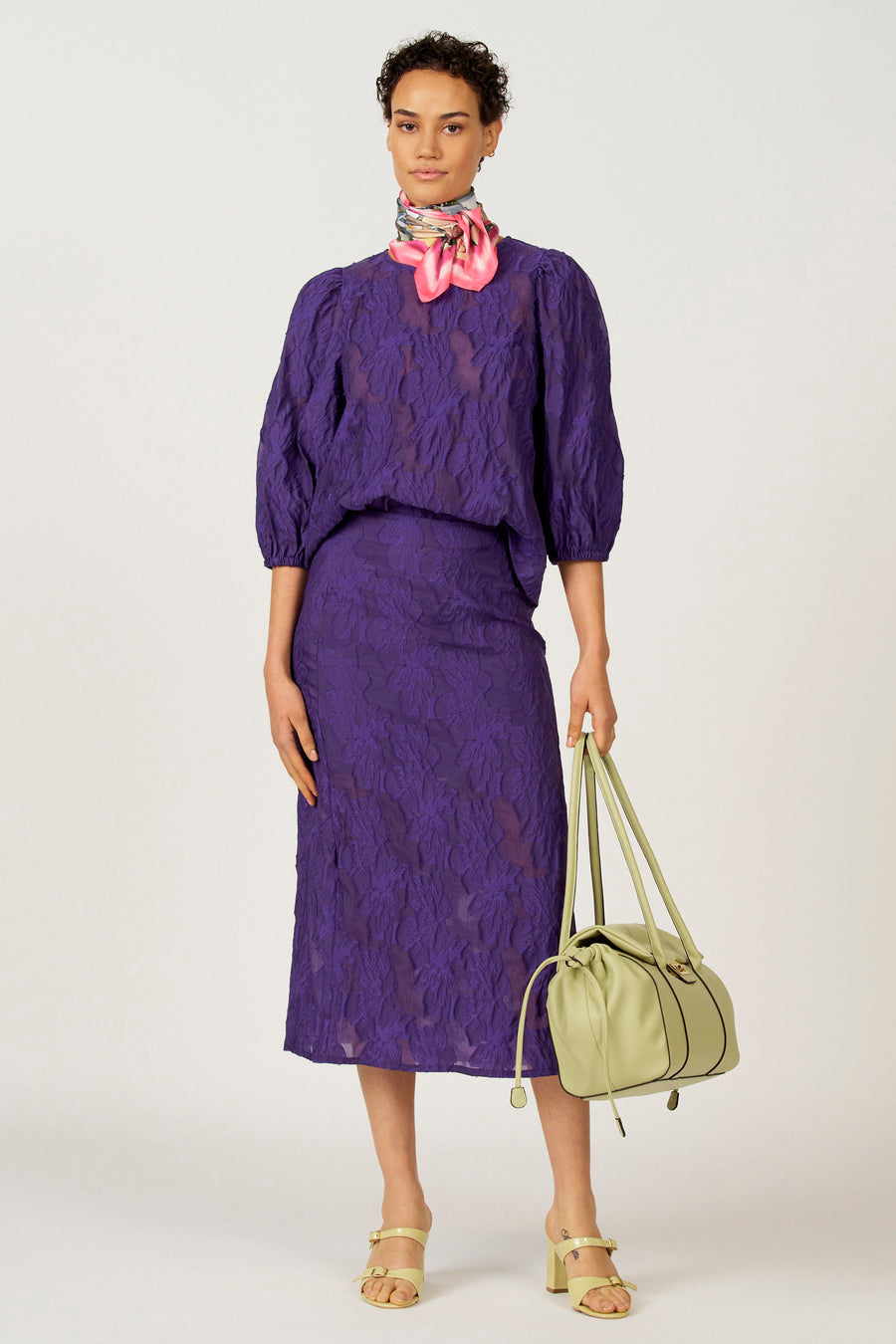 Chemisier violet à manches trois-quarts d'Astrid Black Label, avec un motif floral subtil et combiné avec une écharpe et un sac à main vert.