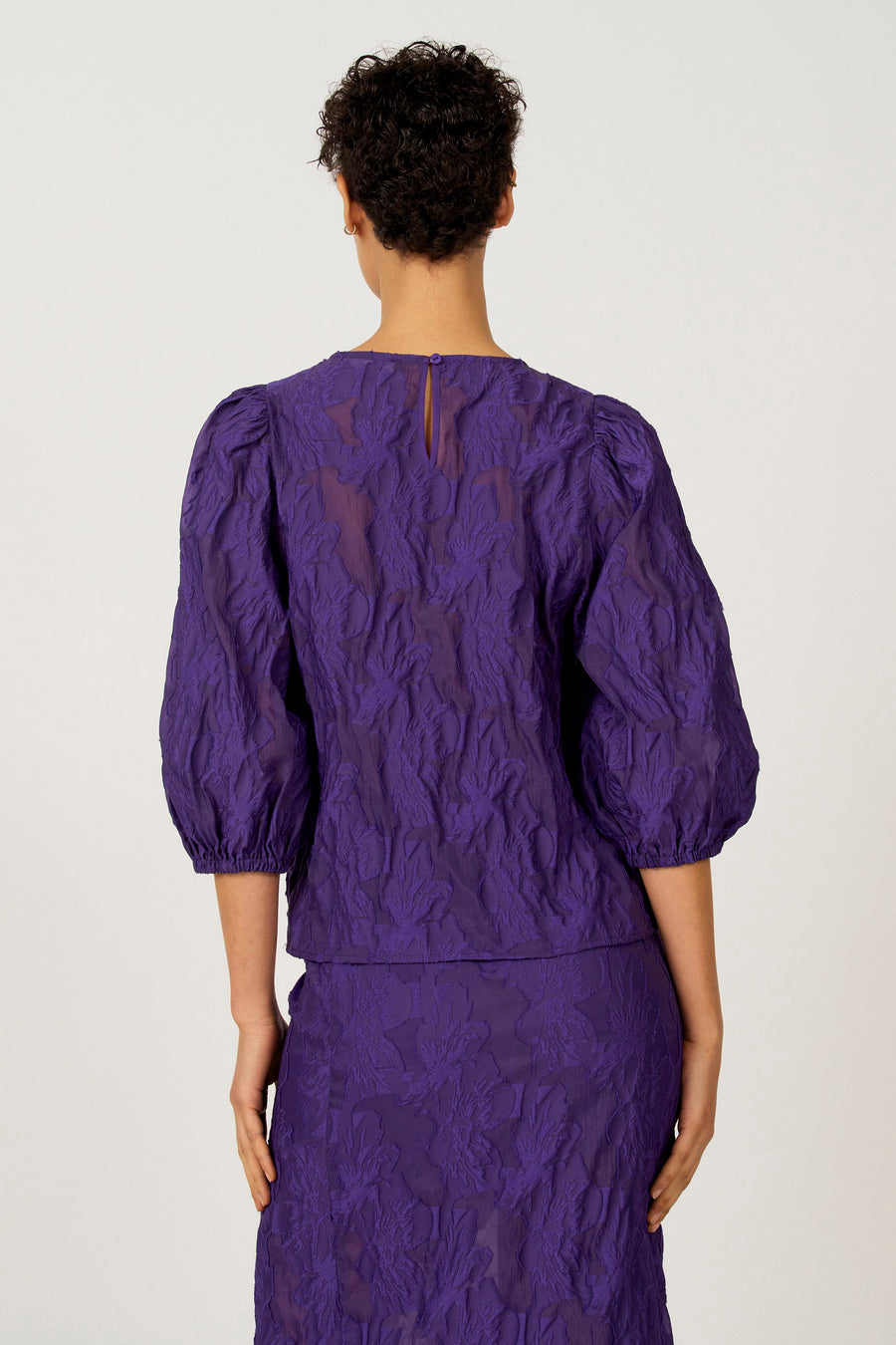 Chemisier violet à manches trois-quarts d'Astrid Black Label, avec un motif floral en relief et une fermeture boutonnée au dos.