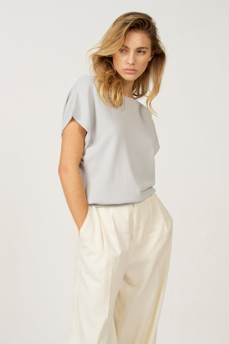 Vrouw met grijze top en crèmekleurige broek, handen in de zakken.