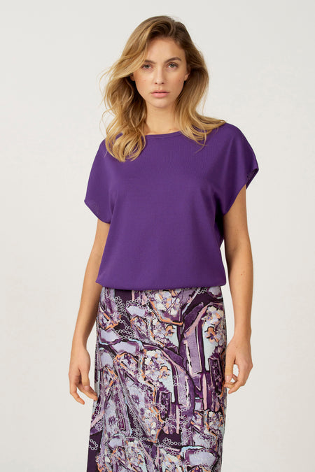 Vrouw in paarse top en paarse rok met print, staand tegen een witte achtergrond.
