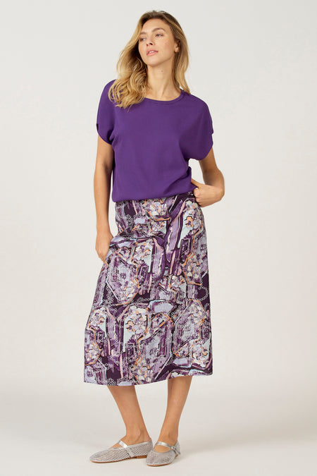 Vrouw in paarse top en rok met print, staand op een witte achtergrond.