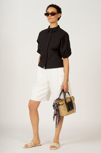 Chemisier noir Astrid Black Label, à manches courtes et porté avec un short blanc et un sac tressé.