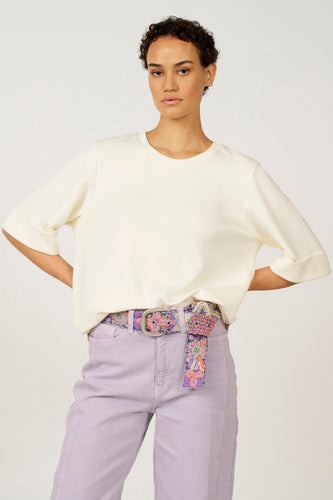 Ecru T-shirt met korte mouwen van Astrid Black Label, met omgeslagen mouwen, gecombineerd met lila broek en riem met bloemenpatroon.
