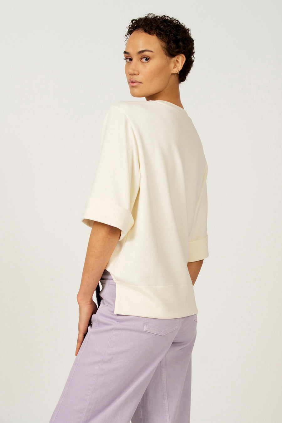 Ecru T-shirt met korte mouwen van Astrid Black Label, met omgeslagen mouwen en zijsplit, gecombineerd met een lila broek.
