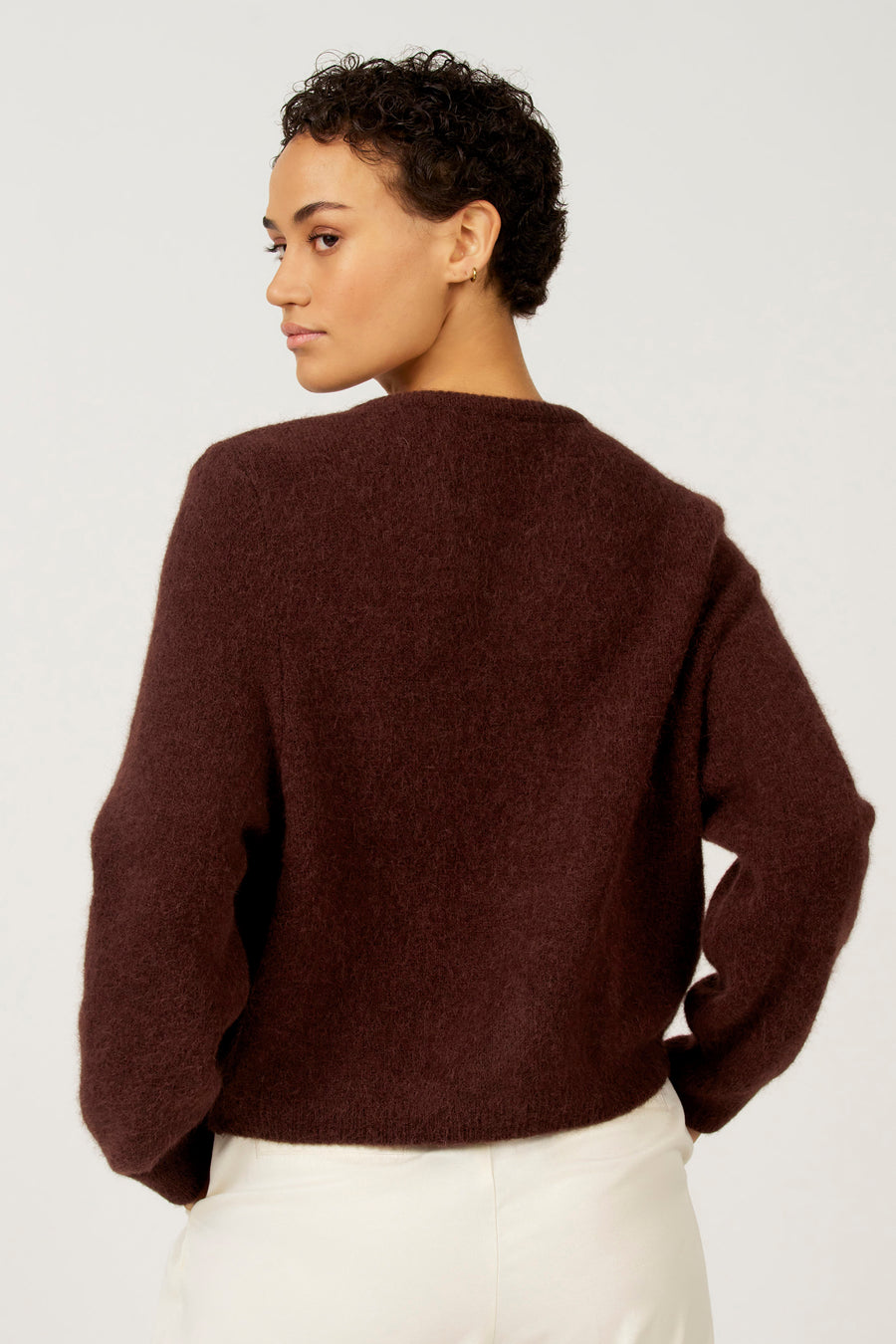 Donkerbruine wollen trui, witte broek, vrouw met kort krullend haar, schuine blik.