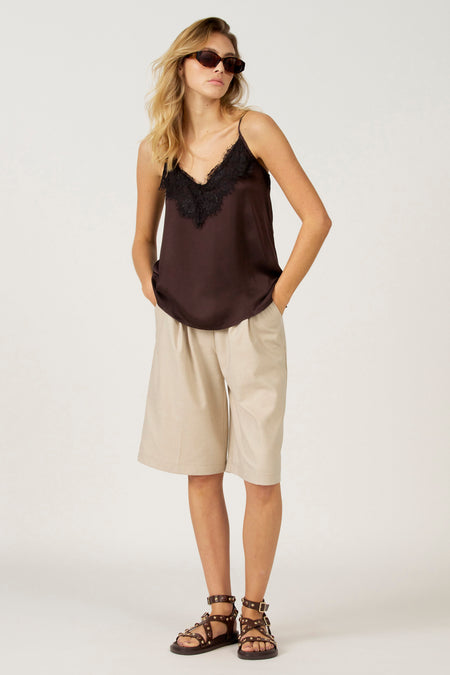 Vrouw in satijnen top met kant, beige shorts en sandalen met noppen, staand.