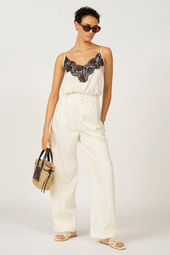 Model draagt witte zijden top met zwart kant, witte broek, zonnebril en een stro tas.