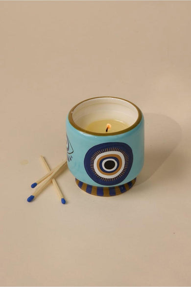 Bougie multicolore de PADDY WAX, avec des motifs oculaires décoratifs bleus et dorés sur un support céramique, à côté de deux allumettes.