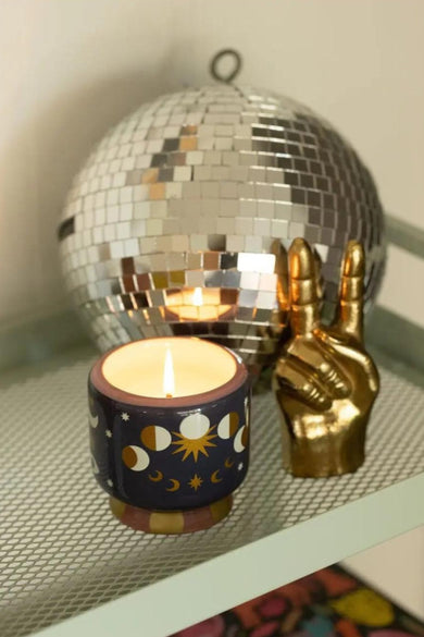 Bougie multicolore de PADDY WAX, avec des phases de lune et un motif d'étoiles, à côté d'un symbole de paix doré et d'une disque.