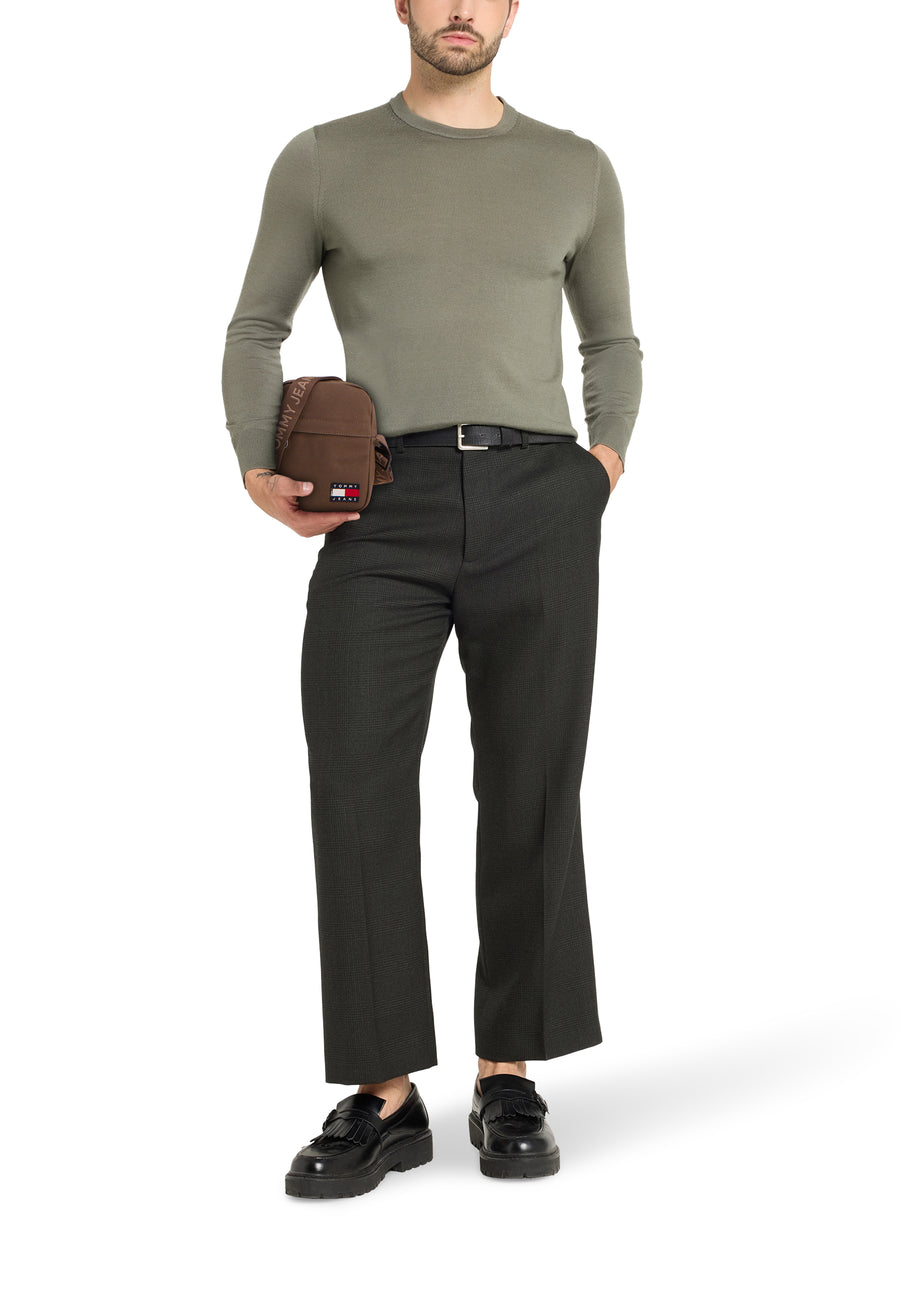 Staande man in groene trui en donkere broek met bruine tas.