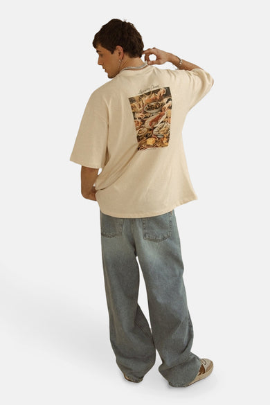 T-shirt écru à manches courtes de KAOTIKO, avec une impression sur le dos et porté avec des jeans larges.