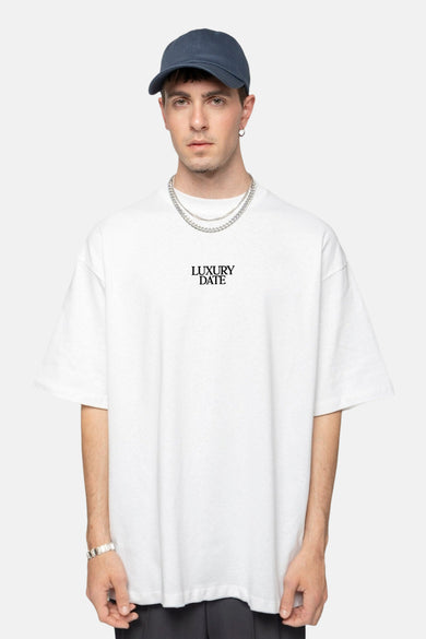 T-shirt blanc à manches courtes de KAOTIKO, avec une impression de texte noire, porté avec une chaîne d'argent et un chapeau.