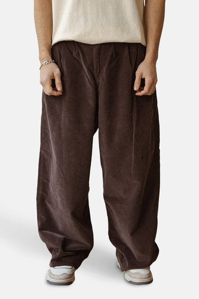 Pantalon brun de KAOTIKO, avec des jambes larges et une légère texture, associé à un shirt crème et à un bracelet argenté.