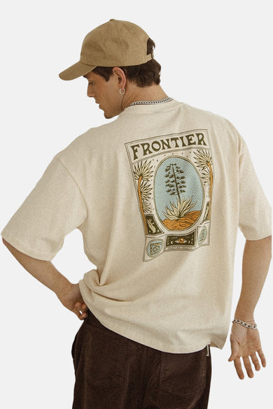 T-shirt écru à manches courtes de KAOTIKO, avec une impression sur le dos, porté avec un pantalon marron et un chapeau.