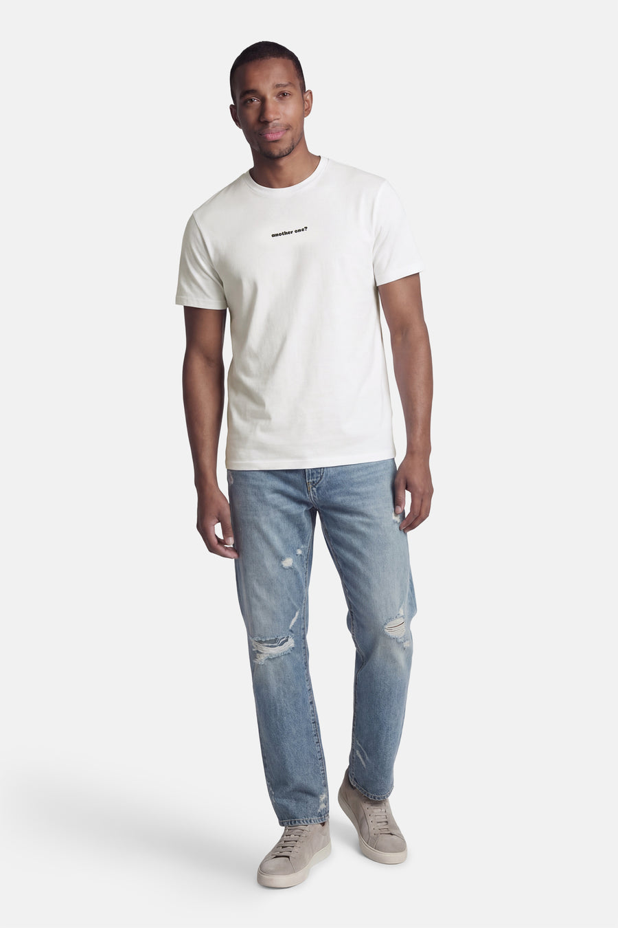 T-shirt blanc de KAOTIKO, à manches courtes et avec un imprimé texte noir, combiné avec un jean bleu déchiré et des chaussures grises.