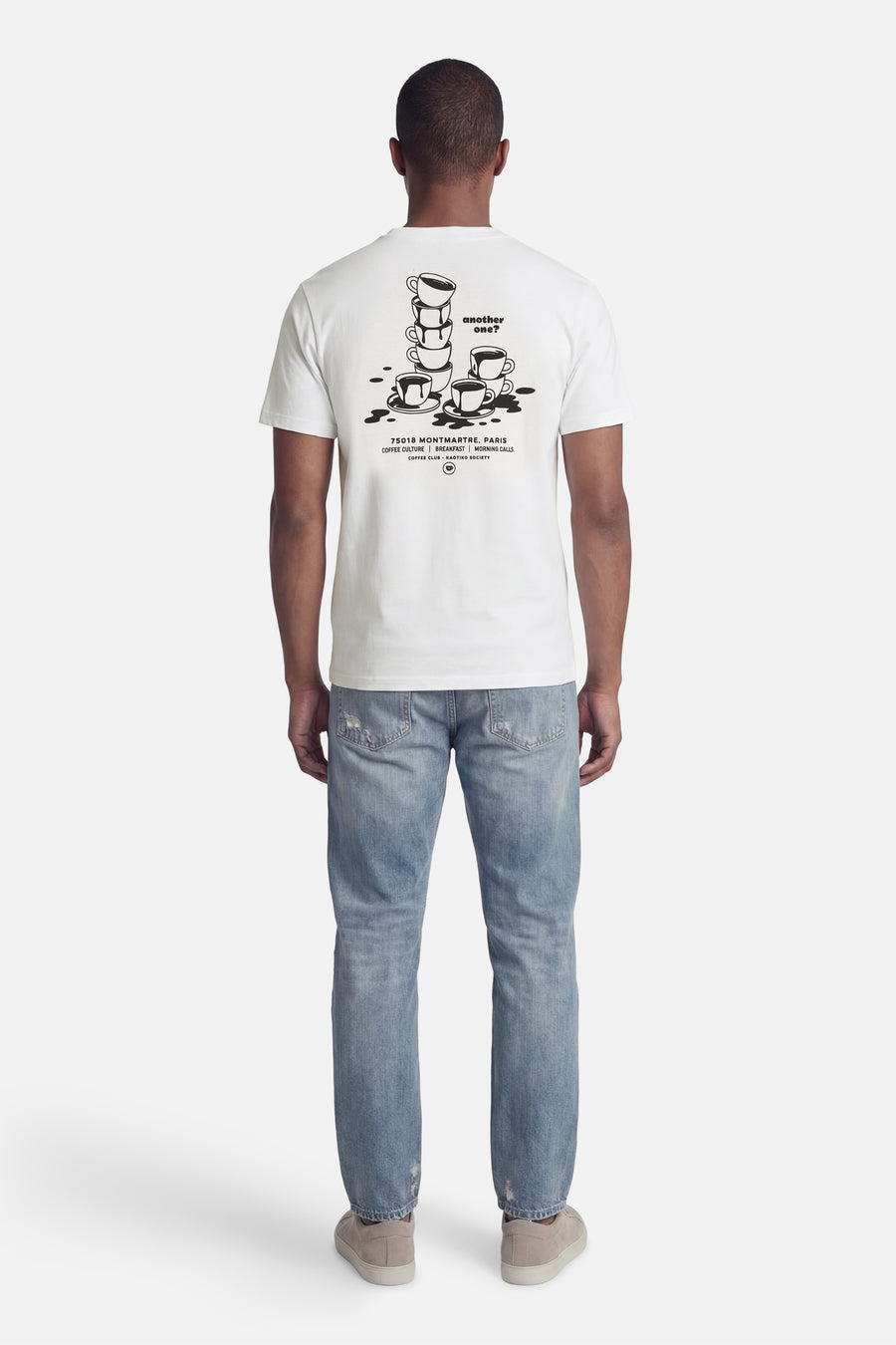 Wit T-shirt van KAOTIKO, met zwarte koffiekopjes print op de achterkant, gecombineerd met blauwe jeans en suède schoenen.

