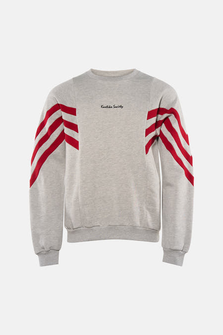 Pull gris de KAOTIKO, avec col rond et rayures rouges sur les manches, combiné avec un jean foncé et des chaussures grises.