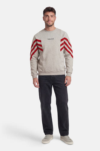 Grijze sweater van KAOTIKO, met ronde hals en rode strepen op de mouwen, gecombineerd met een donkere jeans en grijze schoenen.
