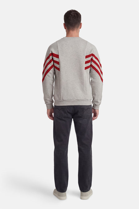 Grijze sweater met ronde hals van KAOTIKO, met rode strepen op de mouwen, gecombineerd met donkere jeans en lichte schoenen, achteraanzicht.
