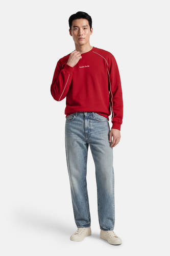Pull rouge à col rond de Kaotiko, fini avec des coutures blanches, combiné avec un jean bleu et des baskets de couleur crème.