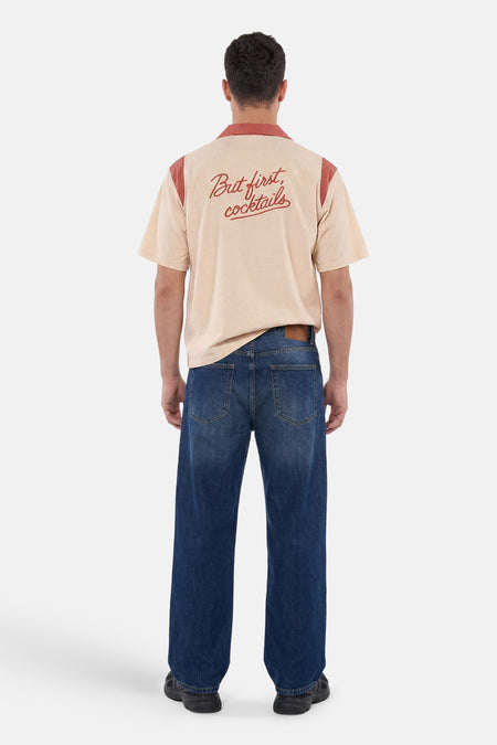 Chemise multicolore à manches courtes de KAOTIKO, beige avec des détails terracotta et 'But first, cocktails' dans le dos, portée par un homme avec un jean foncé.