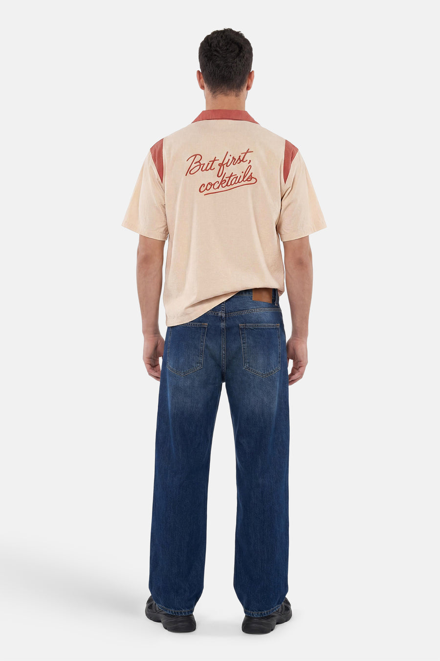 Chemise multicolore à manches courtes de KAOTIKO, beige avec des détails terracotta et 'But first, cocktails' dans le dos, portée par un homme avec un jean foncé.