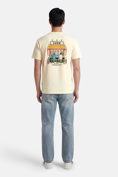 T-shirt écru de KAOTIKO avec un dessin de dessin animé d'un restaurant japonais à l'arrière, porté avec un jean bleu et des baskets blanches.