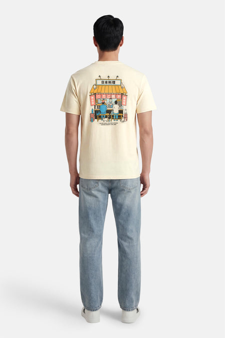 T-shirt écru de KAOTIKO avec un dessin de dessin animé d'un restaurant japonais à l'arrière, porté avec un jean bleu et des baskets blanches.