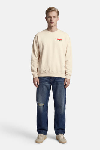 Pull écru à col rond de KAOTIKO, avec imprimé texte rouge sur la poitrine, porté avec un jean bleu déchiré.