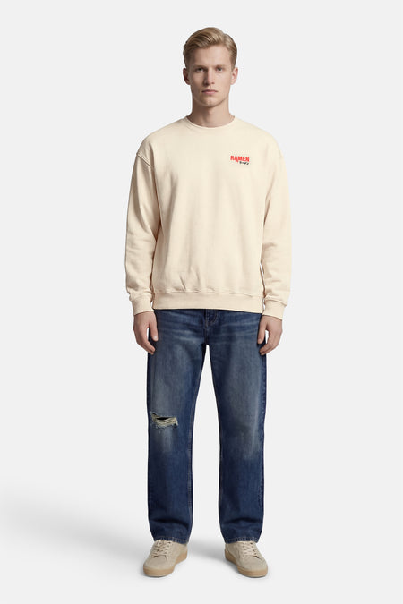 Pull écru à col rond de KAOTIKO, avec imprimé texte rouge sur la poitrine, porté avec un jean bleu déchiré.