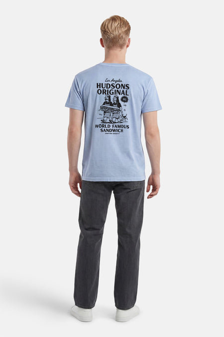 T-shirt bleu clair de KAOTIKO avec un grand imprimé graphique noir au dos de deux personnages portant un sandwich, porté par un homme avec un pantalon gris foncé et des baskets blanches.