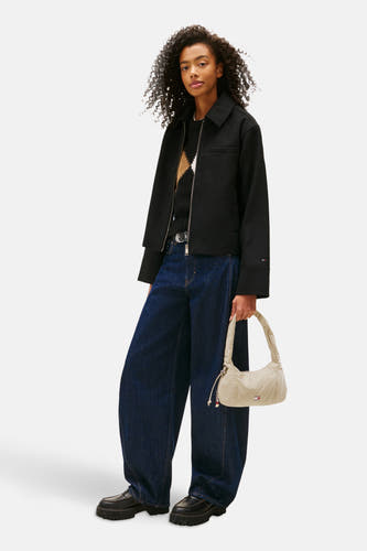 Sac bandoulière beige de TOMMY JEANS, avec fermeture éclair et détails brodés, porté avec un manteau noir et des jeans larges.