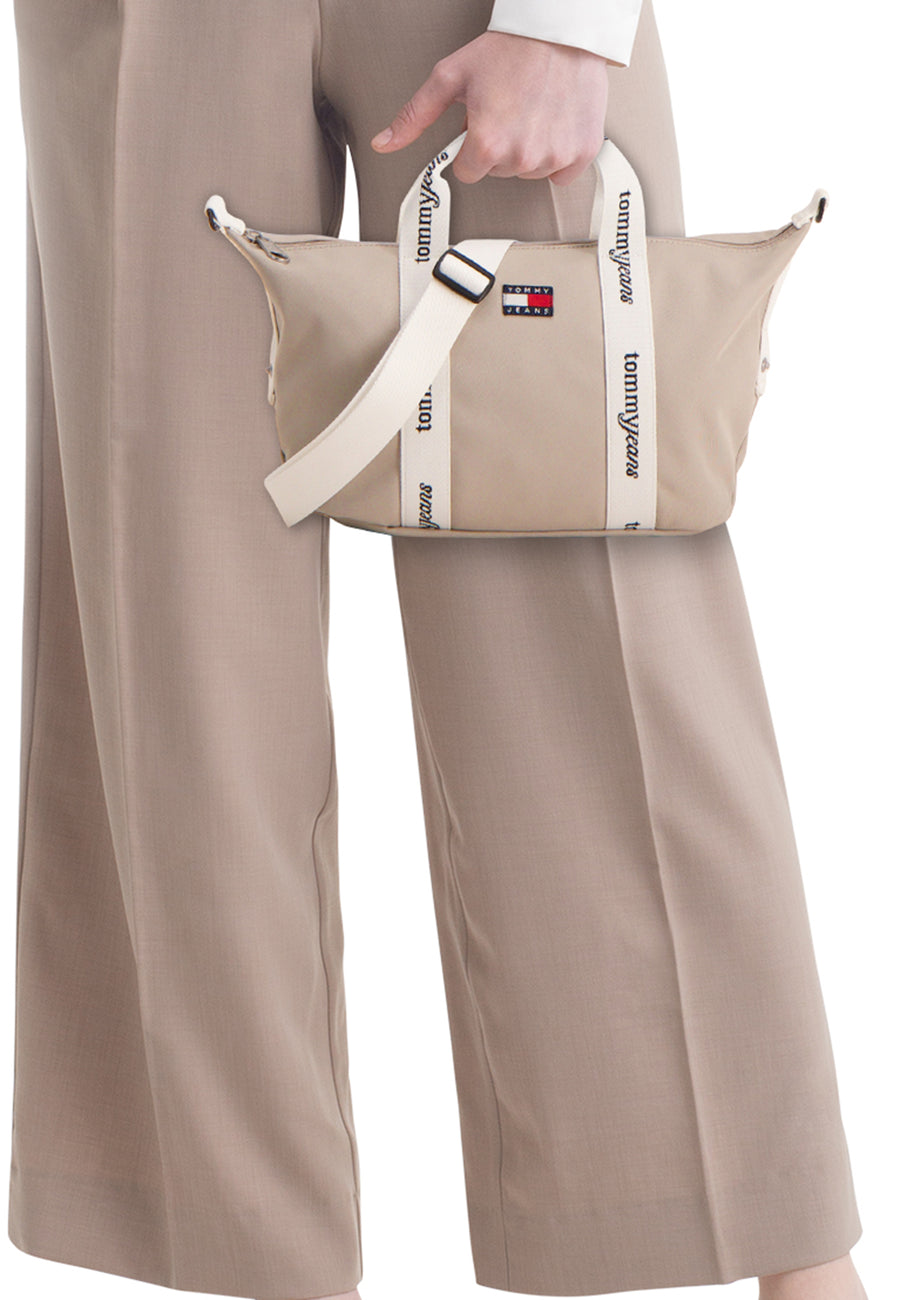 Sac à bandoulière beige de TOMMY JEANS, avec anses blanches à logo, porté avec un pantalon beige.