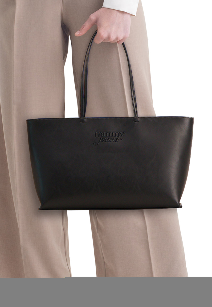 Sac à bandoulière noir TOMMY JEANS avec deux anses et un logo en relief, porté avec un pantalon beige.