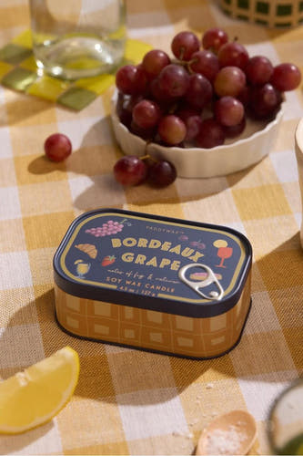 Blauwe kaars in goudkleurig blik met druivenmotief van PADDY WAX, op geruit tafelkleed met fruit en glas water