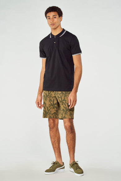 Shorts verts à imprimé palmier de Bruce & Butler, associés à un polo noir et des baskets vertes.