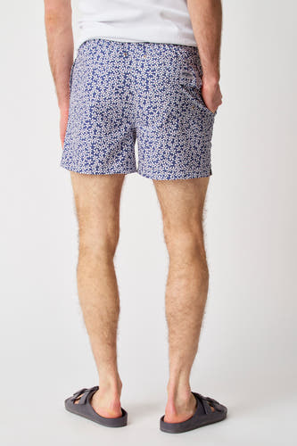 Maillot de bain bleu de Bruce & Butler, avec un motif floral blanc, vu de derrière, porté avec un T-shirt blanc et des chaussons gris.