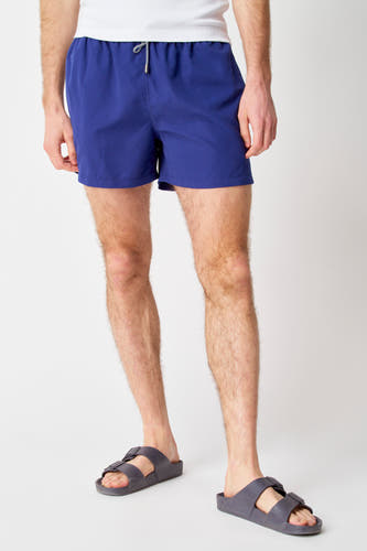 Pantalon de bain bleu Bruce & Butler, porté avec des sandales grises et une chemise blanche.