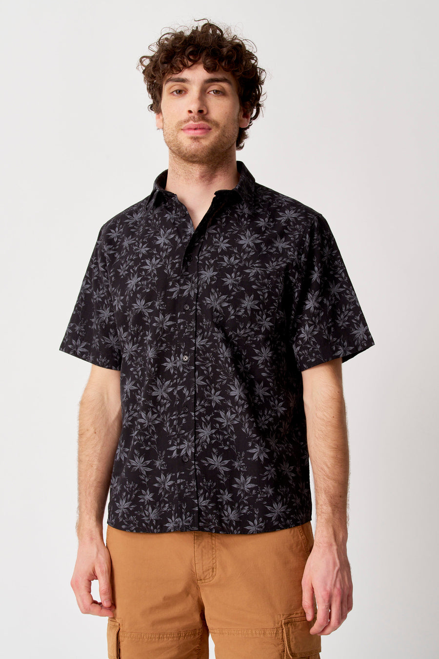 Chemise noire à manches courtes de Bruce & Butler avec motif de feuilles grises, portée par un homme en short cargo marron.