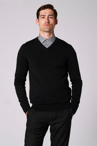 Pull noir à col V de Bruce & Butler, porté avec un chemisier à motif fin dessous, et pantalon noir.