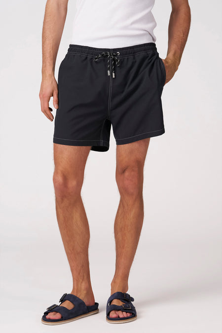 Zwarte zwemshort van Bruce & Butler met wit contraststiksel, trekkoord en zilveren tips, gedragen met wit hemd en blauwe sandalen.