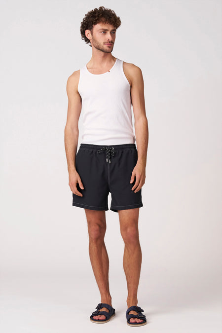 Short de bain noir Bruce & Butler avec surpiqûres contrastées blanches et cordons de serrage rayés, stylisé avec une chemise blanche et des sandales bleu foncé.