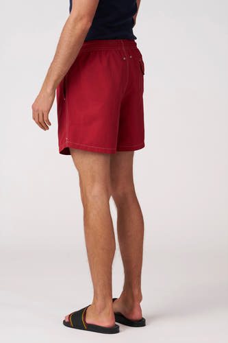 Short de bain rouge de Bruce & Butler avec surpiqûres blanches, taille élastique, poches latérales et arrière, porté avec une chemise bleu foncé et des sandales noires, rouges et jaunes, vu de dos et de côté.