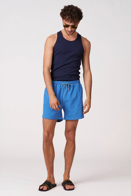 Short de bain bleu de Bruce & Butler avec cordon de serrage et coutures blanches, porté par un homme avec un débardeur bleu marine, des lunettes de soleil et des tongs noires.