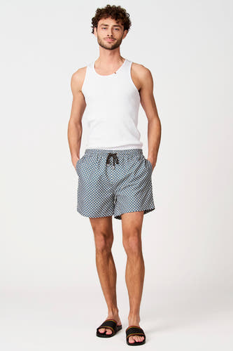 Short de bain bleu-vert à motif géométrique blanc de Bruce & Butler, porté par un homme avec une chemise blanche et des sandales rayées.